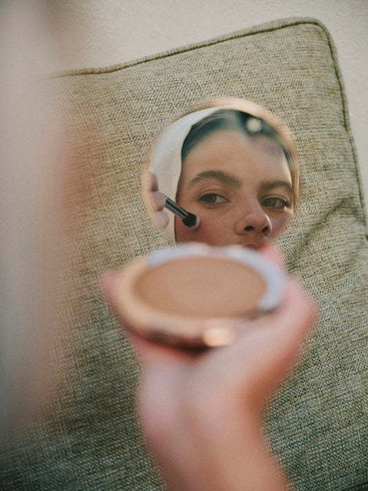 Person applying powder makeup using a brush, reflected in a small round mirror