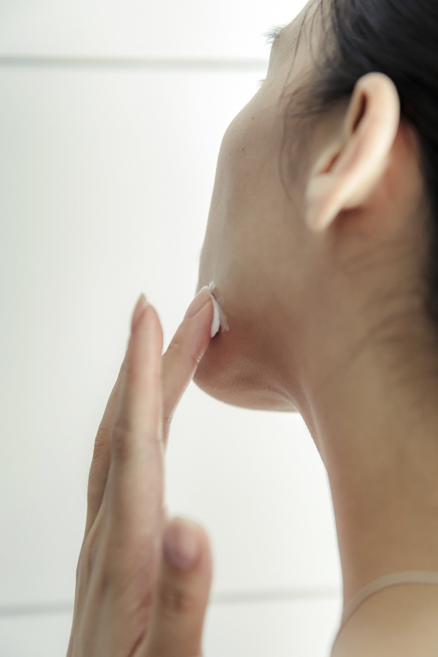 A person applies a dab of white cream to their jawline with one finger, shown in profile against a plain background.