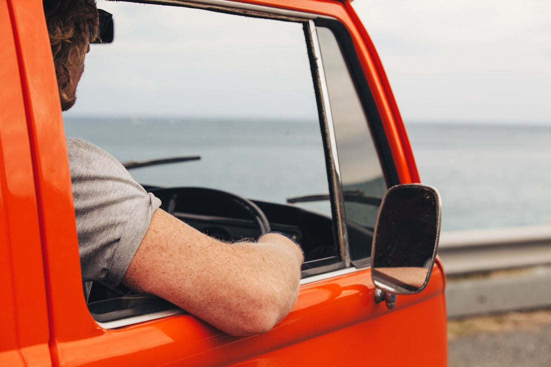 Person driving with arm resting on an open car window, ocean visible in the background.