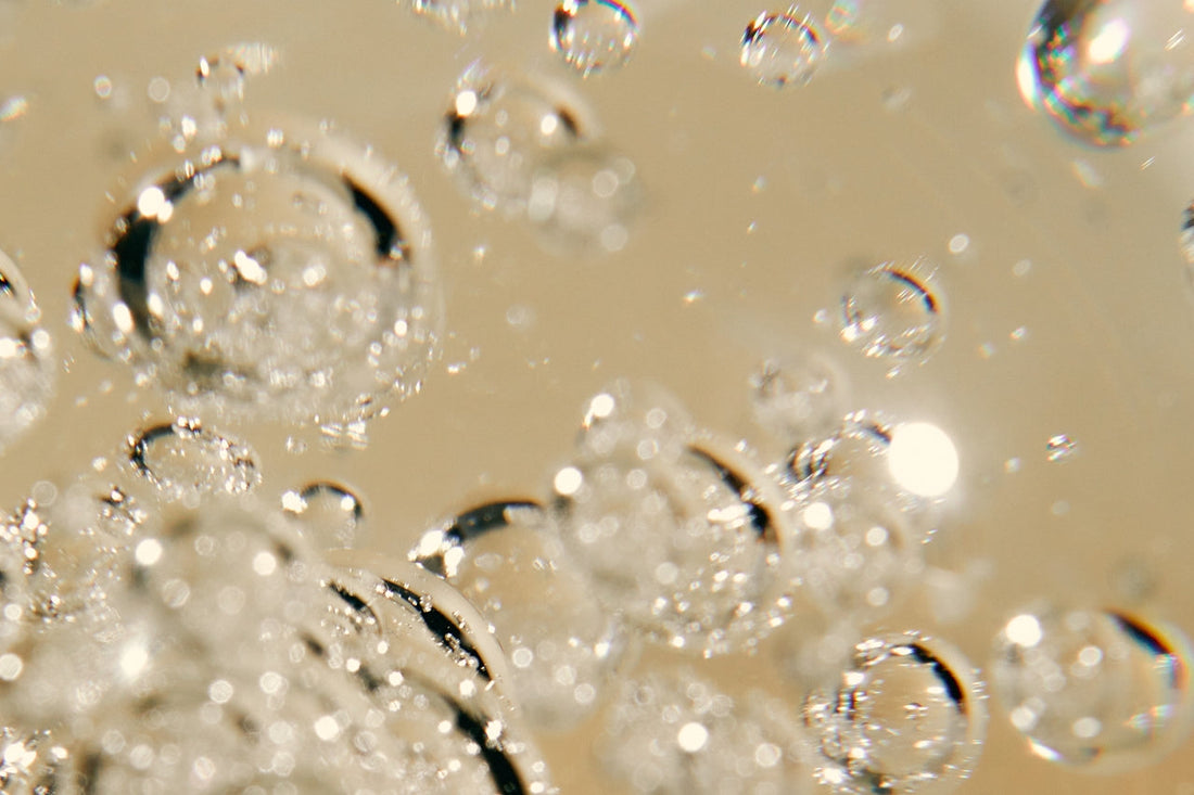 Macro view of clear air bubbles suspended in a golden liquid, with light reflecting off their rounded surfaces against a warm beige background.