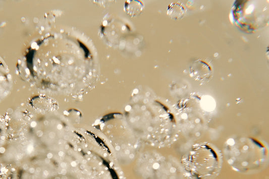 Macro view of clear air bubbles suspended in a golden liquid, with light reflecting off their rounded surfaces against a warm beige background.