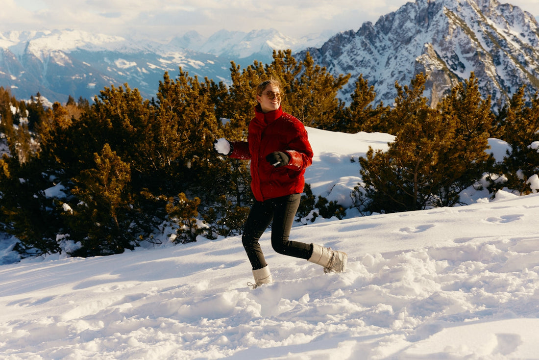 A person in a red jacket tosses a snowball while running across a snowy mountain trail, with evergreen trees and snow-covered peaks in the background.