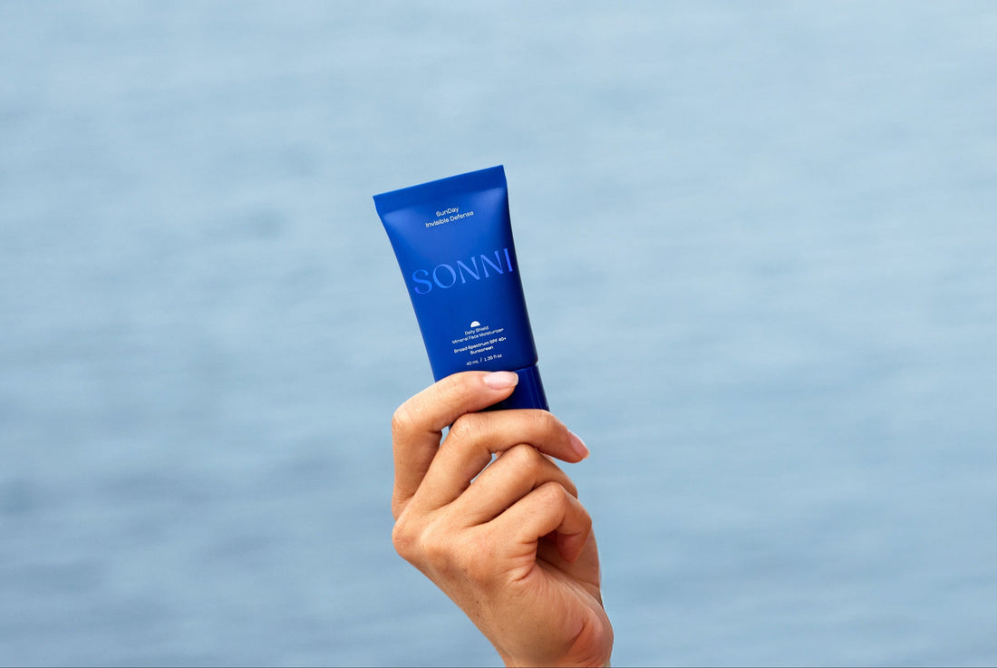 Hand holding a blue SONNI mineral suncare tube against a soft blue sky background.